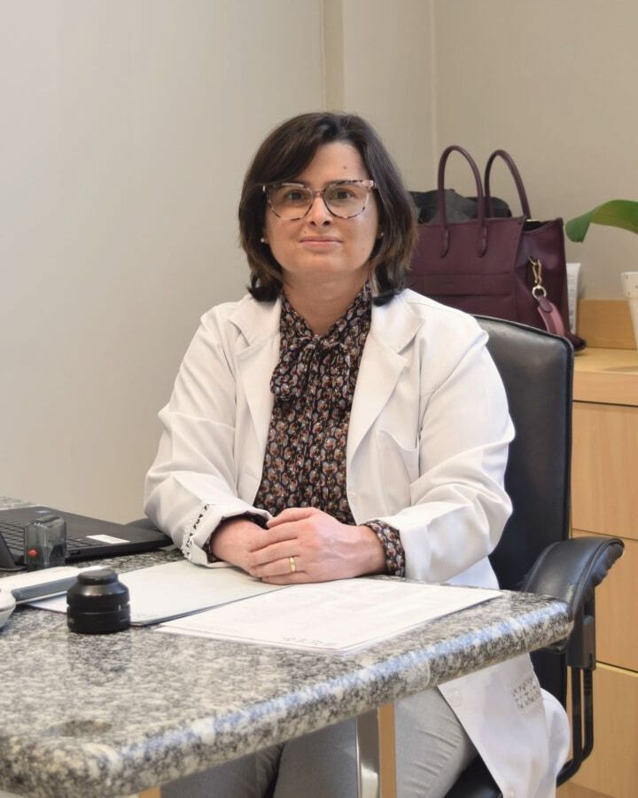 Dra. Daniela Gontijo, dermatologista, de jaleco branco, sentada à mesa do consultório na Mooca, em São Paulo